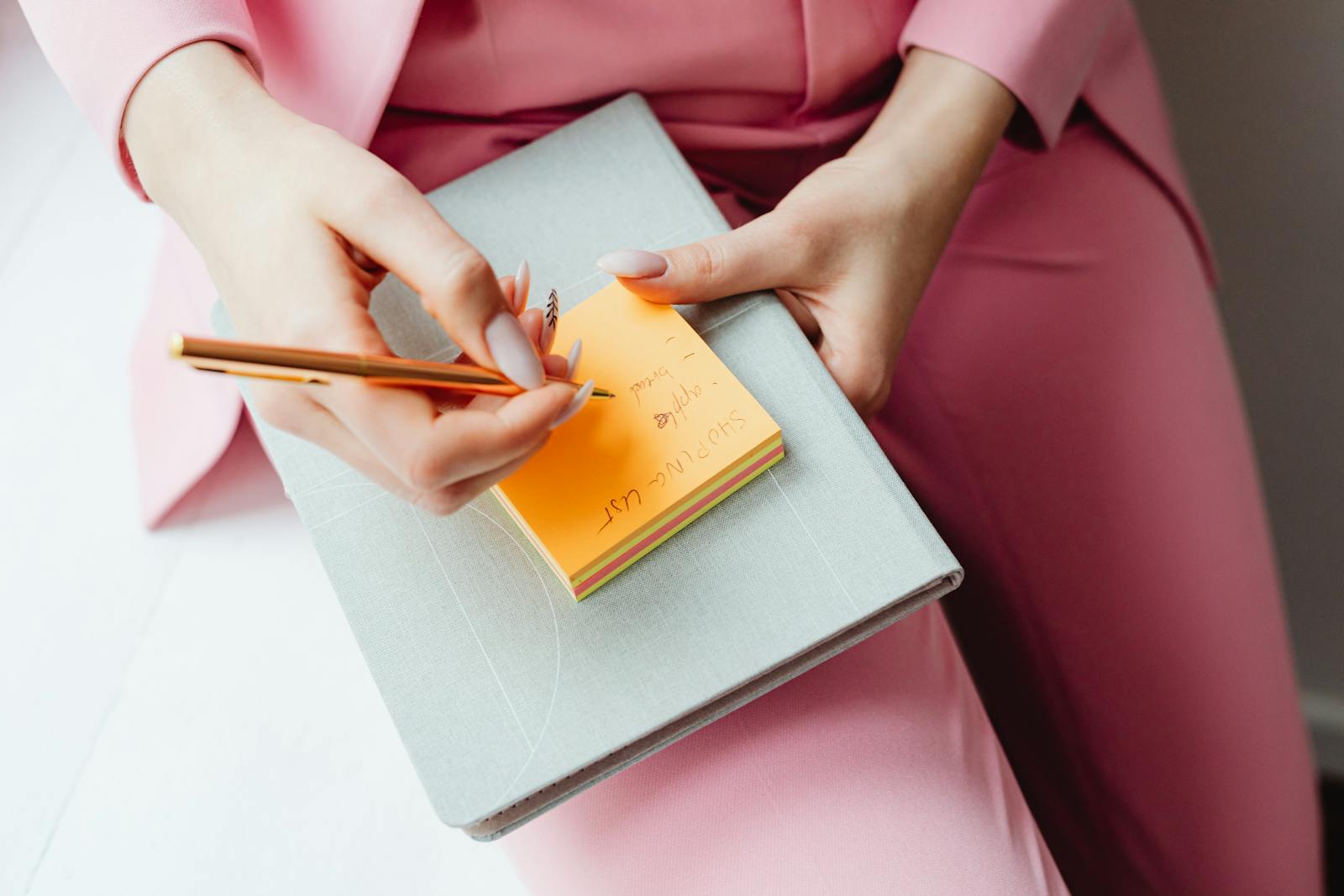 Close-up of a woman writing on a sticky note with a pencil, wearing a pink suit.