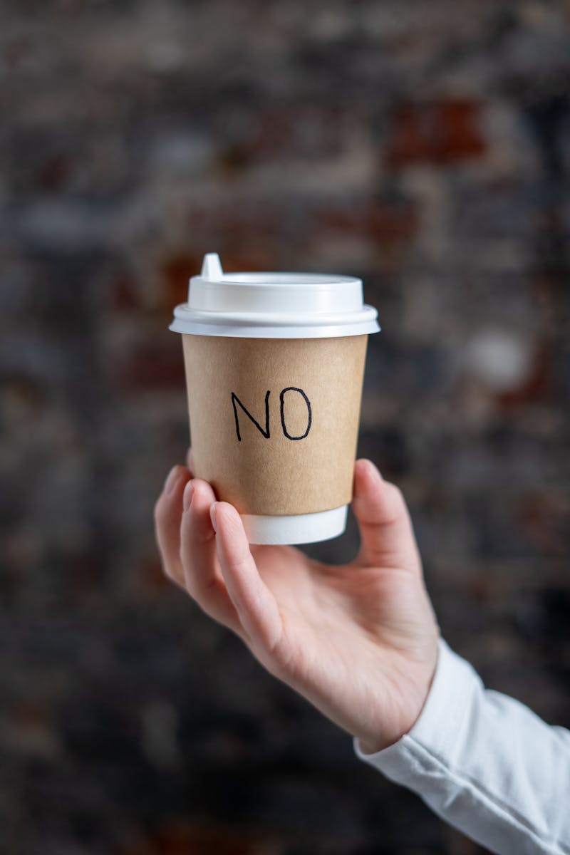 A hand holding a paper coffee cup with the word NO written on it, against a blurred background.