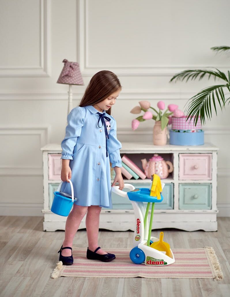 A young girl in a blue dress happily plays with a toy cleaning set inside a stylish room.