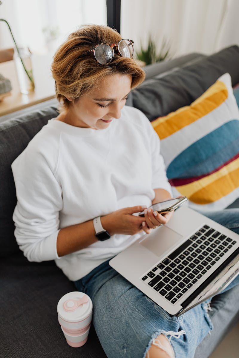 Woman in white sweater using phone and laptop on couch at home office.