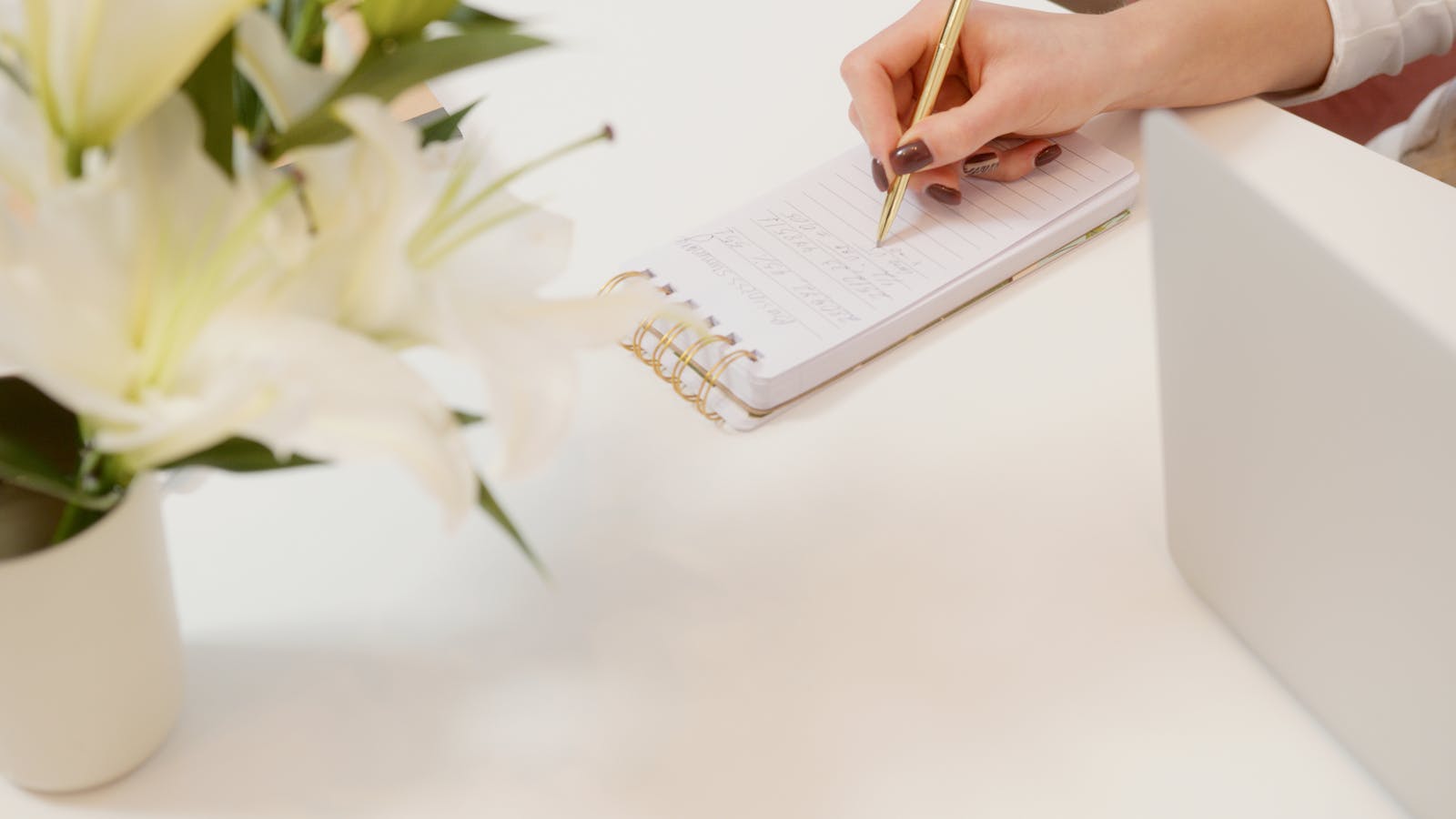 A workspace with lilies, notepad, and gold pen on a white desk.