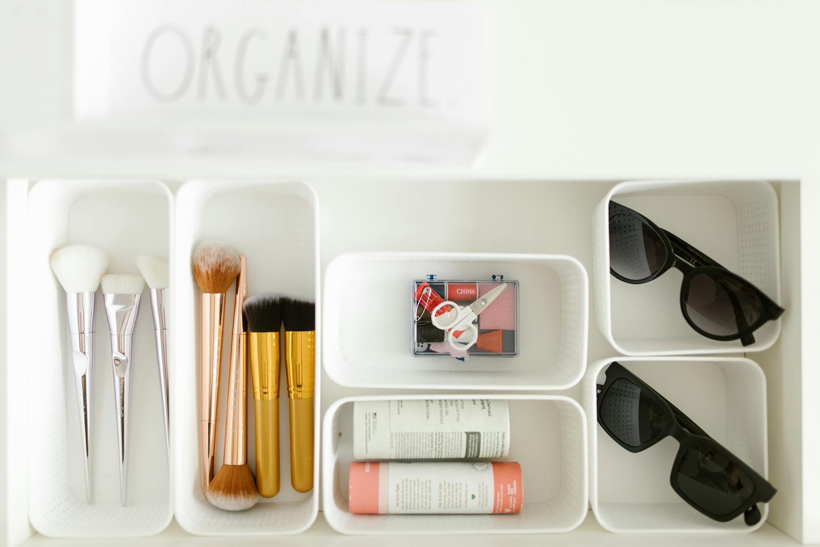 Flat lay of cosmetics and accessories neatly organized in white trays on a white background.