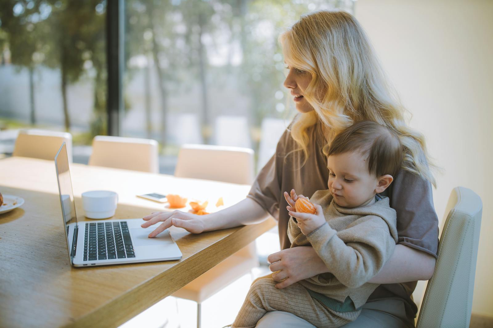 A mother works on a laptop at home while holding her baby, showcasing remote working and parenting.