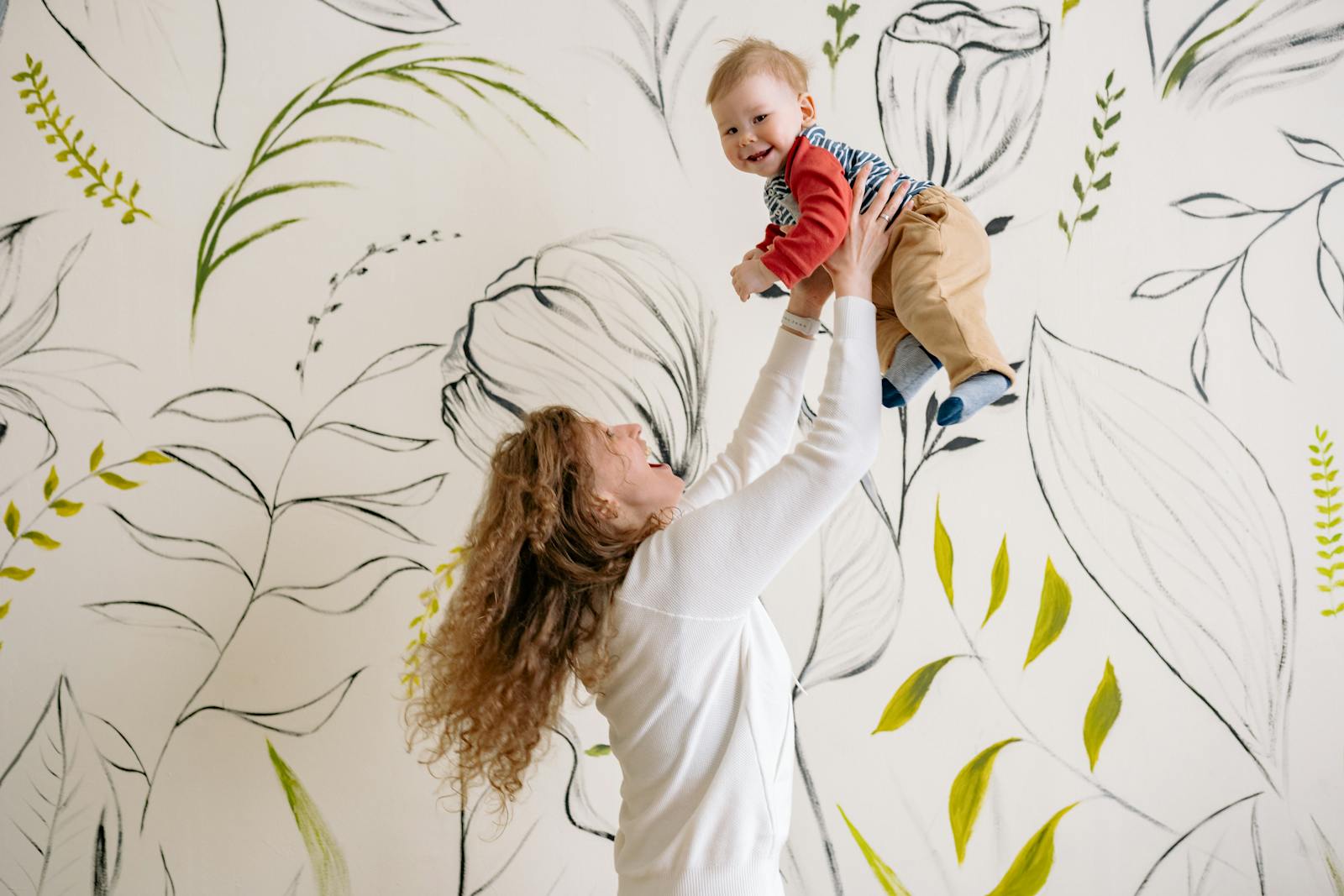 Mother joyfully lifts her smiling baby indoors with a beautiful floral background.